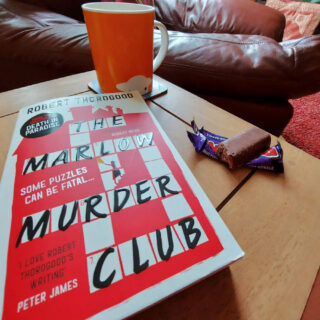 A copy of a book called "The Marlow Murder Club" is lying on a wooden table next to an orange mug of tea and a chocolate bar with a bite out of it. In the background is a brown leather chair.
