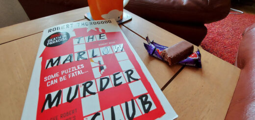 A copy of a book called "The Marlow Murder Club" is lying on a wooden table next to an orange mug of tea and a chocolate bar with a bite out of it. In the background is a brown leather chair.