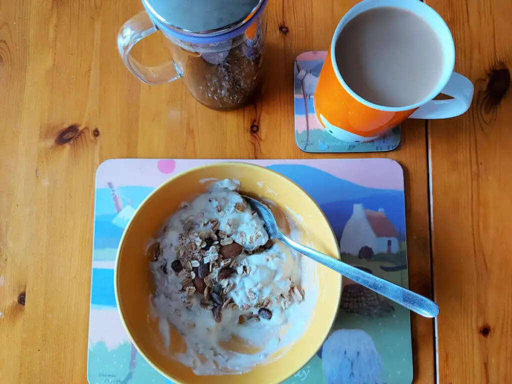 A yellow bowl of muesli and yoghurt sits on a brightly coloured placemat on a wooden table. To the top right of the mat is a smaller version of the same mat with an orange mug of tea on it, and to the left is an empty tea pot.