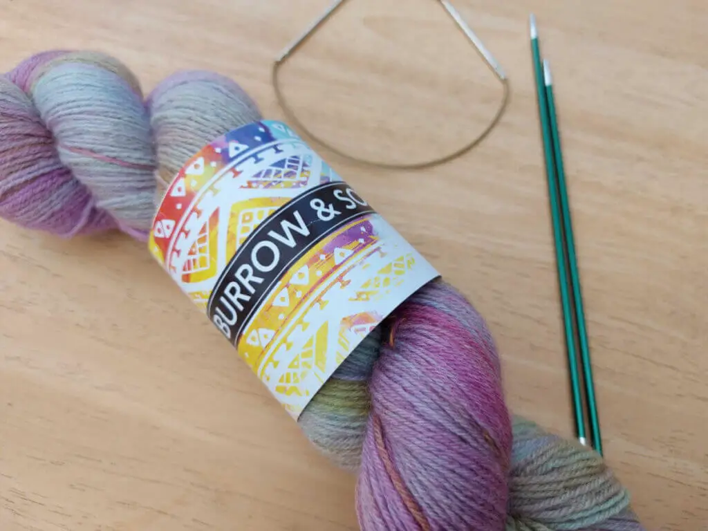 A skein of yarn in soft pastel colours lying on a wooden table with a short circular needle and two green double pointed needles.