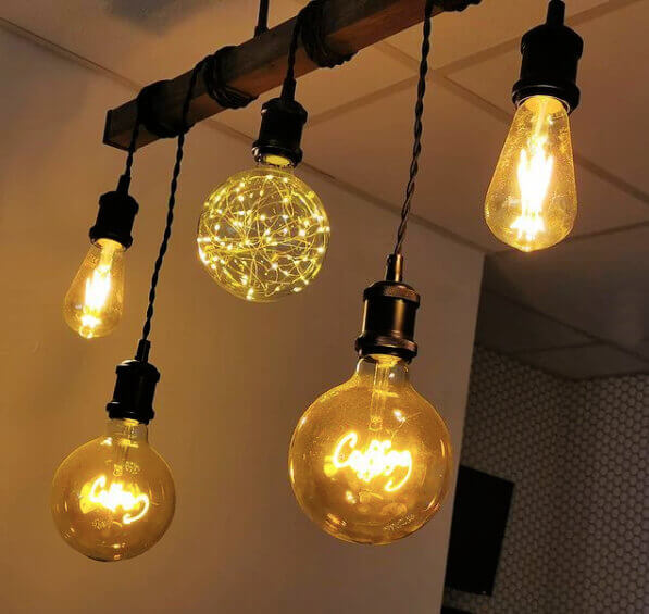 Five ornate lightbulbs of different sizes hanging from a wooden bar. Two of the lights have the word 