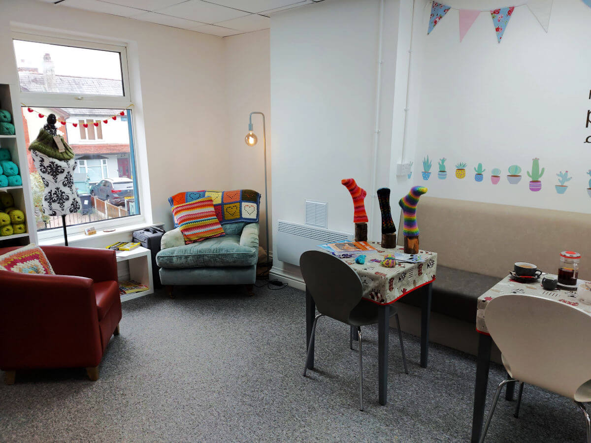A red leather club chair, a pale blue armchair and tables and chairs are arranged along the wall opposite the yarn shelves.