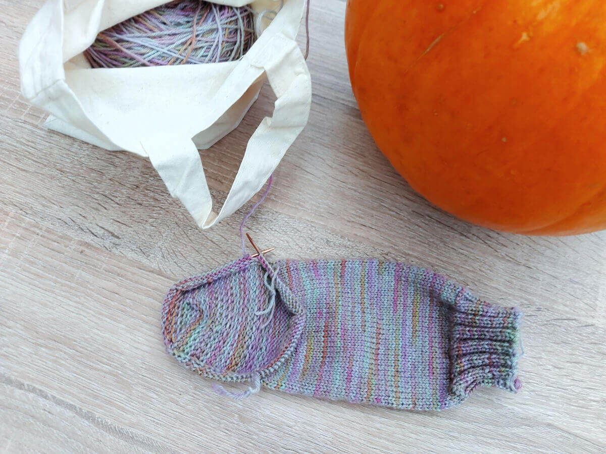 A half-knitted sock on a table next to a bag containing the yarn and a pumpkin.