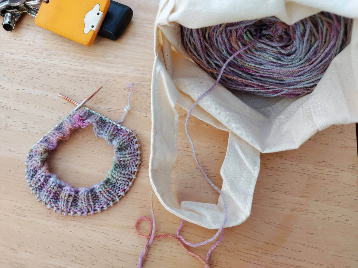 The start of a sock cuff is on a short circular needle and the rest of the pastel yarn cake is inside a cream fabric bag with handles. To the top left are car keys with an orange Herdy key ring.
