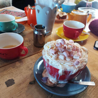 A pine table is littered with large coloured cups and saucers, milk jugs, and knitting projects. In the foreground is a cup of hot chocolate overflowing with cream and marshmallows