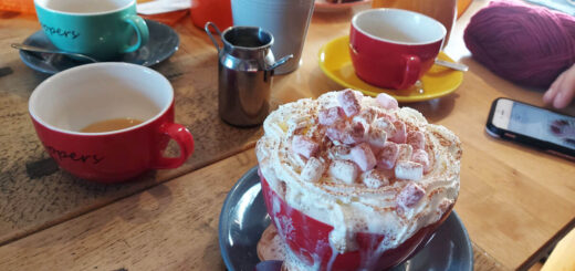 A pine table is littered with large coloured cups and saucers, milk jugs, and knitting projects. In the foreground is a cup of hot chocolate overflowing with cream and marshmallows