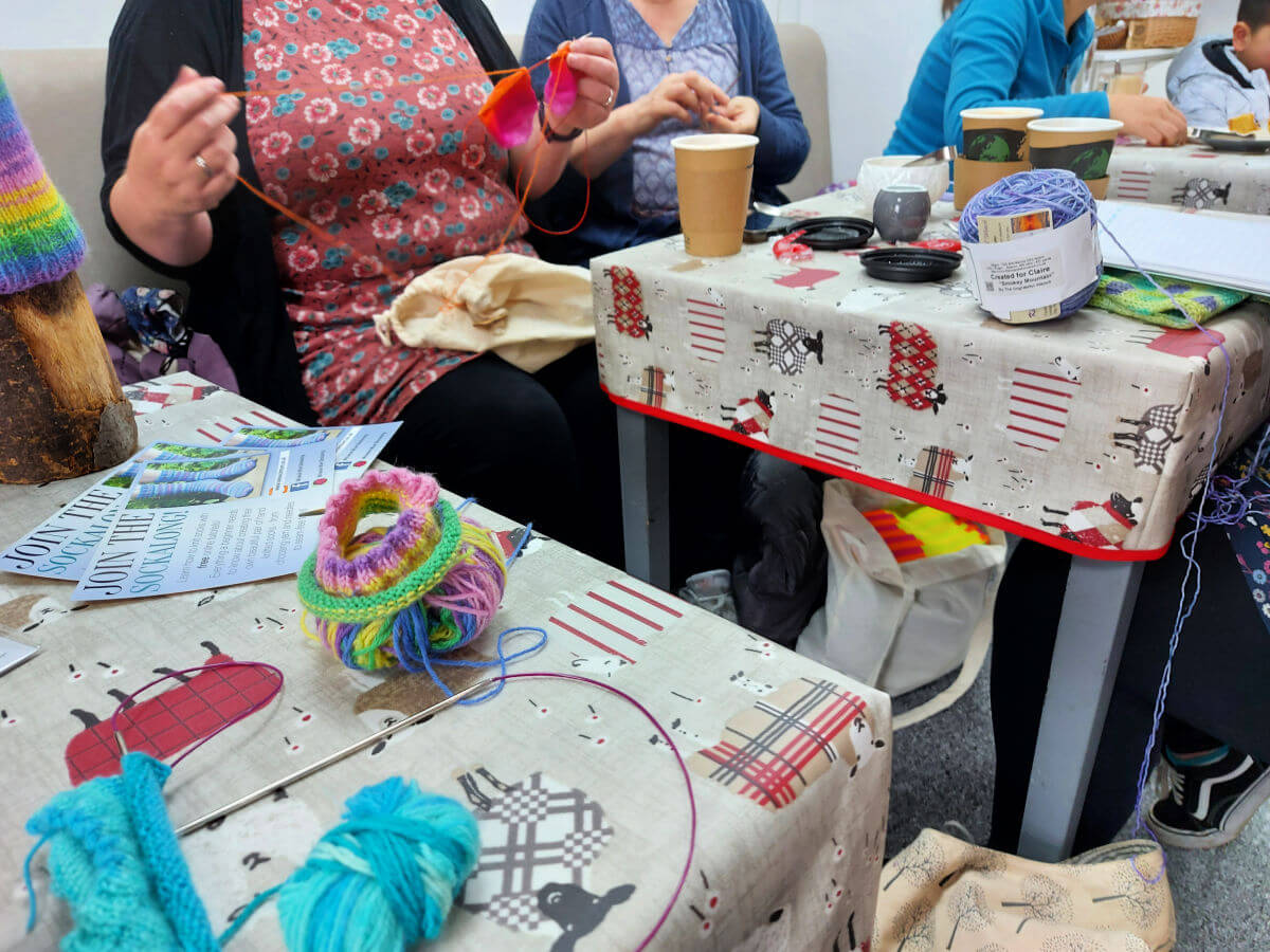 The coffee tables are filled with yarn, projects and coffee cups, and there are people sitting around them working on the projects.