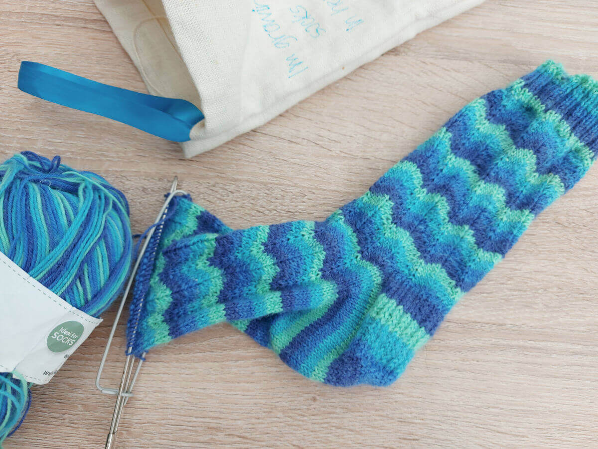 A striped sock in a ripple pattern is almost finished and the stitches are on holders.  The rest of the ball of yarn and the project back are next to the sock.