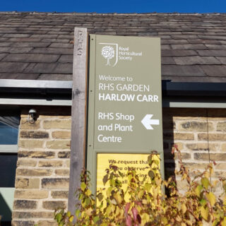A sign at RHS Harlow Carr against a stone building and a blue sky.