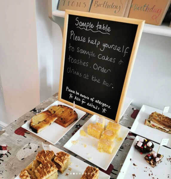A blackboard stands on table filled with plates of cake cut into bite-sized pieces.
