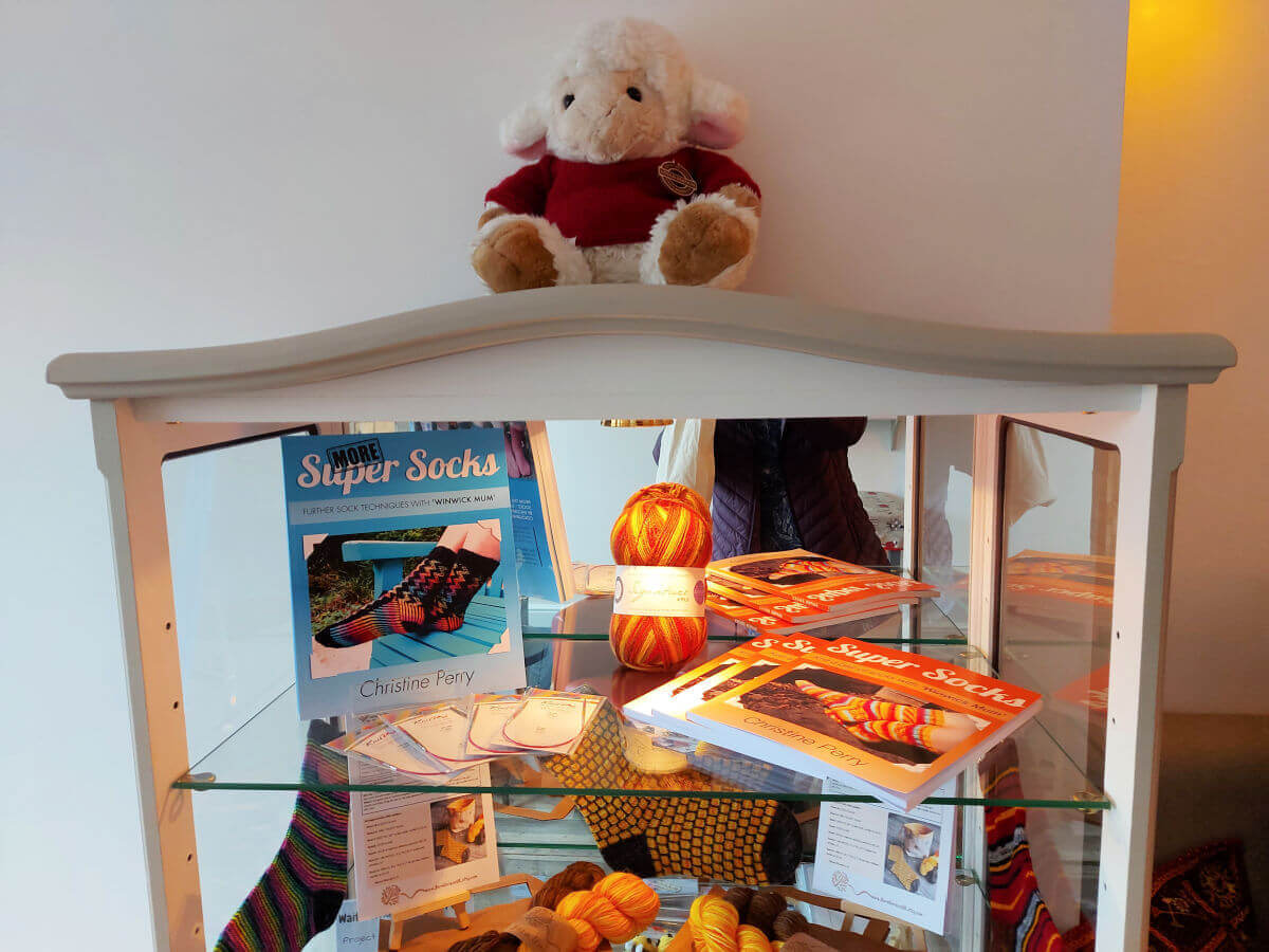 Winwick Mum books, balls of yarn and knitting needles on the top shelf of the showcase.