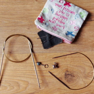 A large circular needle, a short circular needle, two stitch markers and a small fabric purse with wool needles are on a wooden floor.