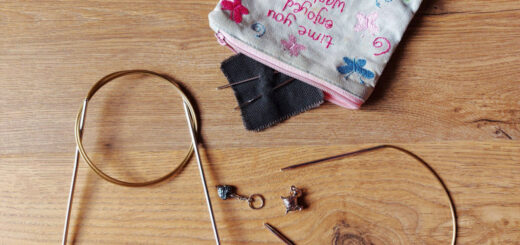A large circular needle, a short circular needle, two stitch markers and a small fabric purse with wool needles are on a wooden floor.