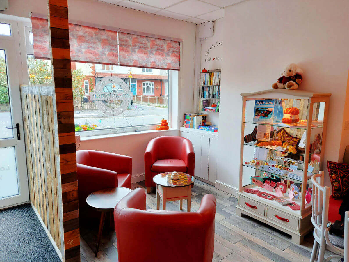Comfy red chairs around a small coffee table near the window. There is a cabinet of local craft items to the right and on the wall, a book case for borrowing and exchanging books.