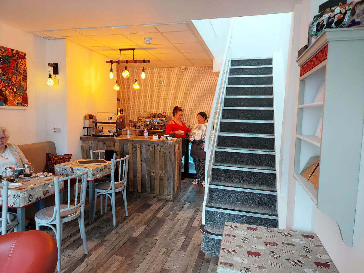 A cosy and welcoming cafe with a barista station at the far end of the room, tables and chairs to the left and stairs on the right which lead to the upper floor.