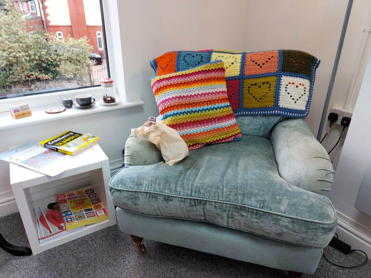 A large pale blue velvet armchair sits in a corner with a crocheted blanket and cushion on it. To the left is a small cabinet with crochet patterns on it, and above that is a window. 