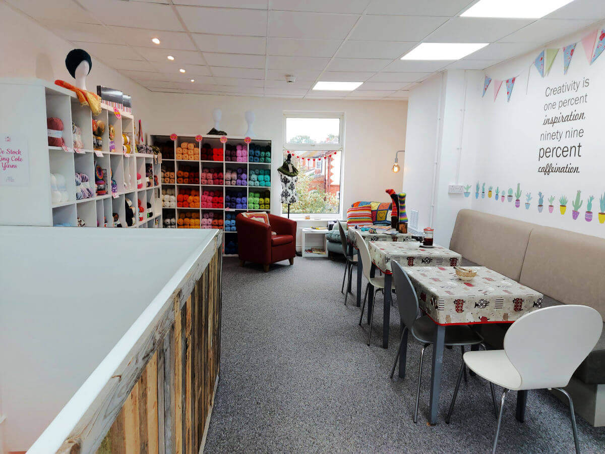 A view of the upper floor of the Stitch Station. There are yarn shelves to the left after the staircase opening, tables and chairs to the right and comfy armchairs at the end by the window. The room is light and bright.