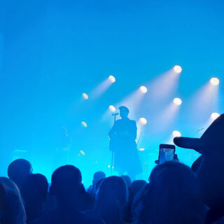 A man is standing on a stage backlit with blue lights. In the foreground are lots of heads of the crowd.