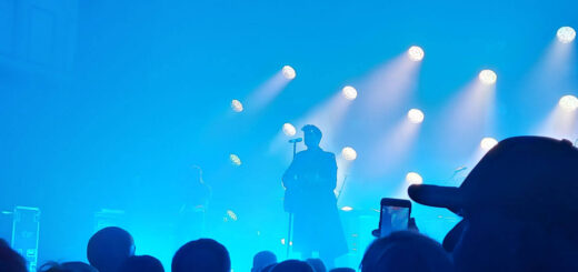 A man is standing on a stage backlit with blue lights. In the foreground are lots of heads of the crowd.
