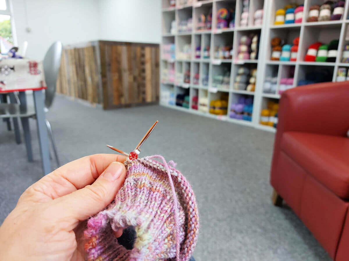 Christine is holding a partly-knitted sock on a short circular needle and looking down the length of the Stitch Station upper floor. There are yarn shelves to the right and chairs and tables to the left.