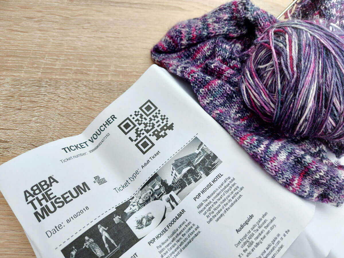 Printed tickets to the ABBA museum in Stockholm lie on a table next to a partly-knitted purple mottled sock.