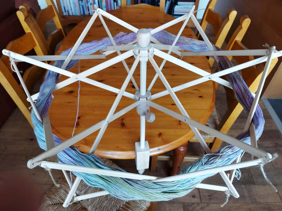 A skein of pastel coloured yarn opened up and draped around a wooden umbrella swift. The ribs of the swift hold the yarn safely whilst it turns. The swift is attached to a pine dining table. There are chairs around the table and bookcases in the background.