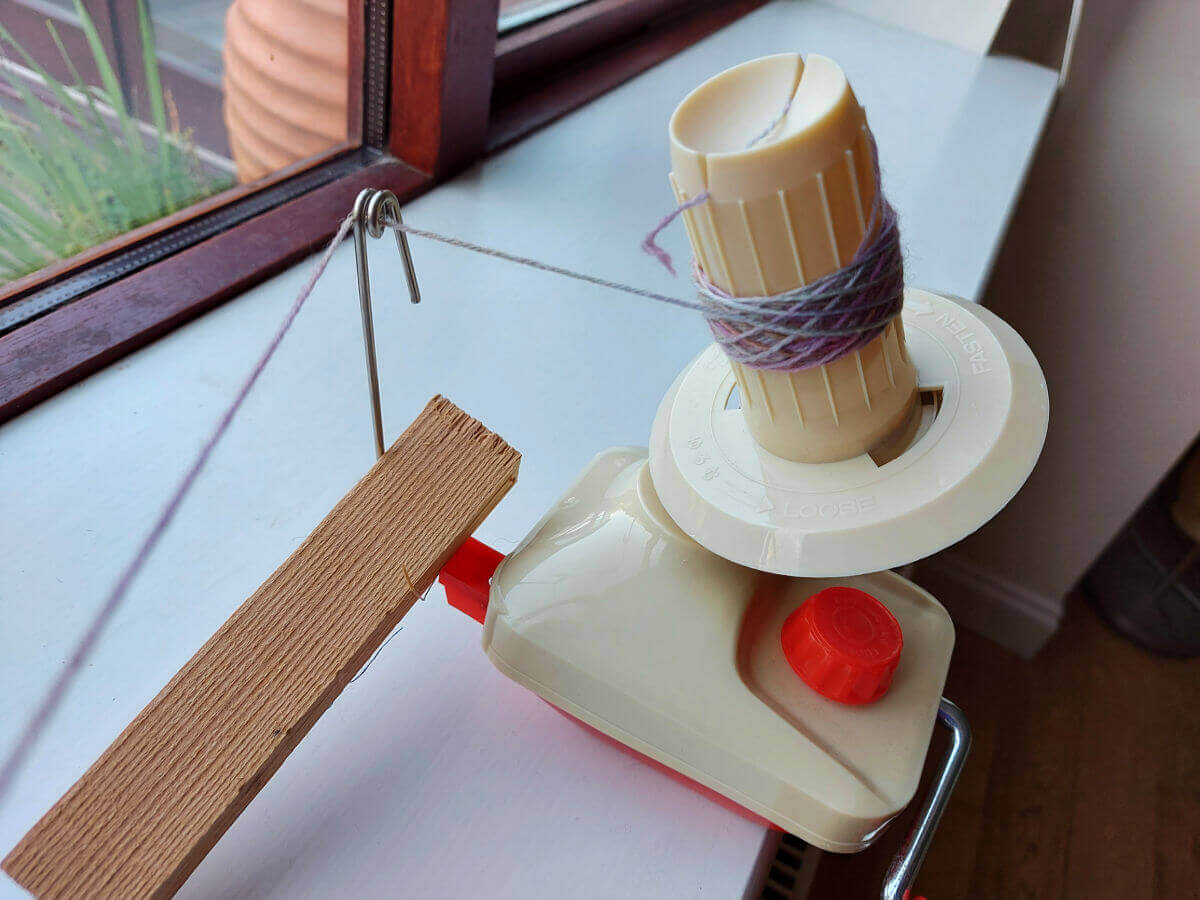 A red and cream yarn winder attached to a window sill. A piece of wood stops the guide falling over and there is a small amount of yarn wound onto it.