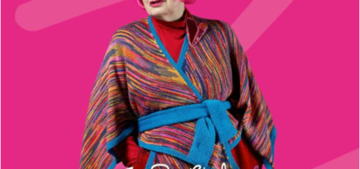 Dame Zandra Rhodes stands against a pink background that matches the colour of her hair. She is wearing a hand knitted wrap cardigan in shades of pink and blue.