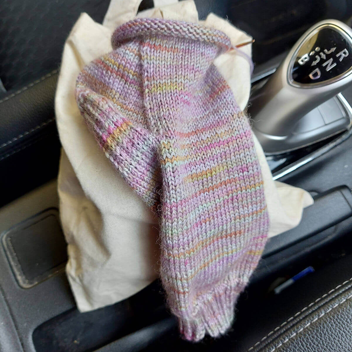 A partly-knitted sock sits on a cream project bag next to a car gear stick.