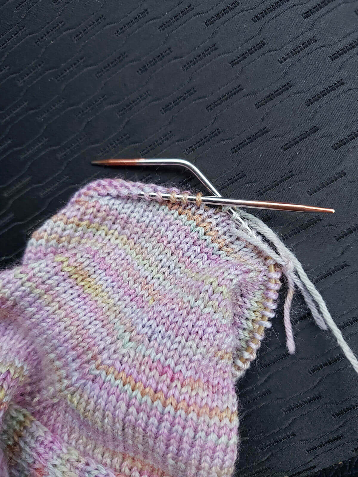 A partly-knitted sock on a short circular needle resting on a black car seat. One of the needle tips is bent at nearly 90 degrees.