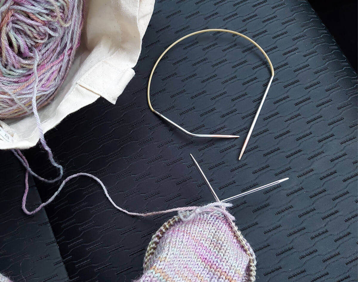 A partly-knitted pastel-coloured sock on a short circular needle.  Both the needle tips are straight.  There is a short circular needle with one bent tip lying on the black car seat next to it, and there is a cream fabric project bag containing the yarn in the top left of the photo.