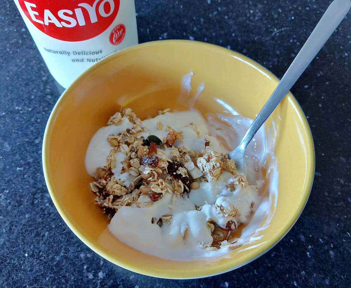 A yellow dessert bowl containing muesli and natural yoghurt is sitting on a granite worktop. There is a stainless steel spoon in the bowl. Next to the bowl is an Easiyo yoghurt pot.