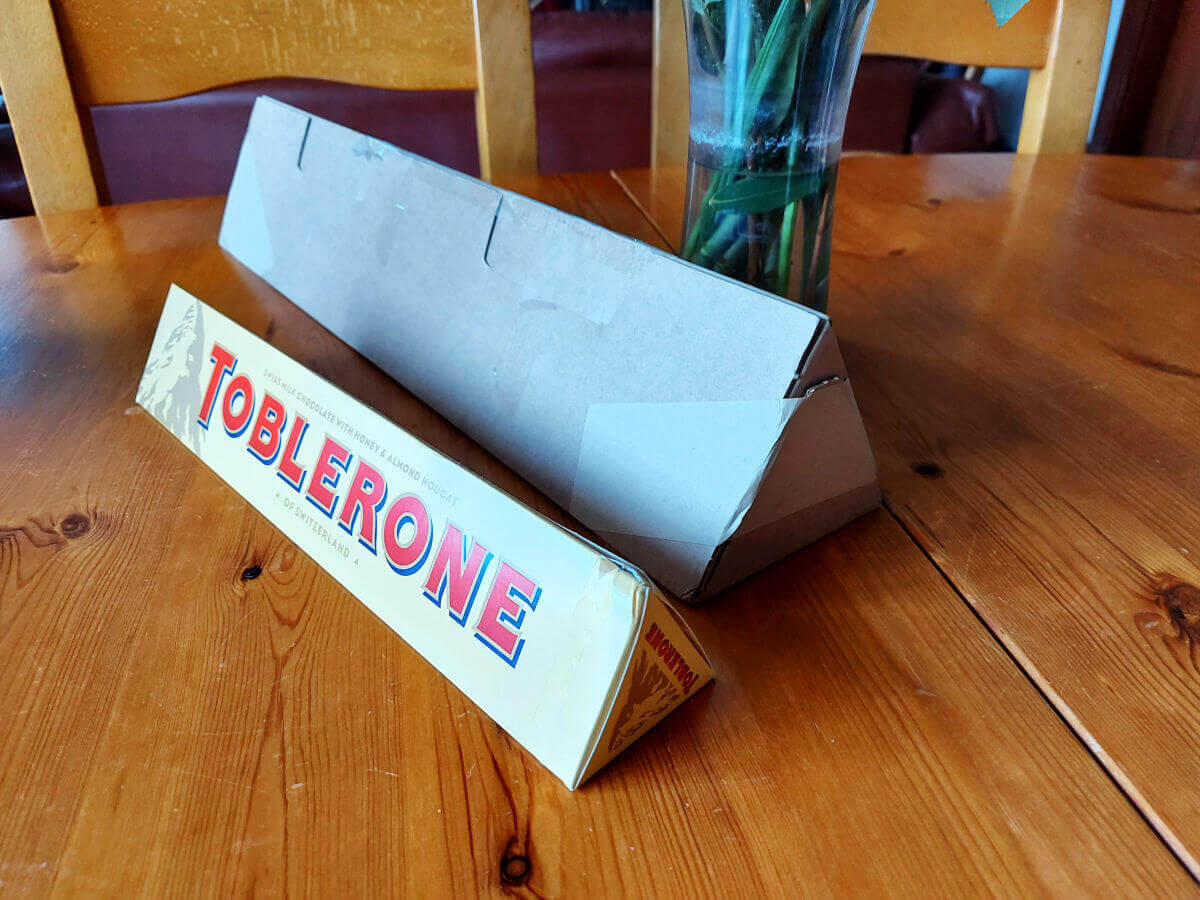 A very large triangular cardboard box on a wooden table next to a smaller Toblerone triangular box