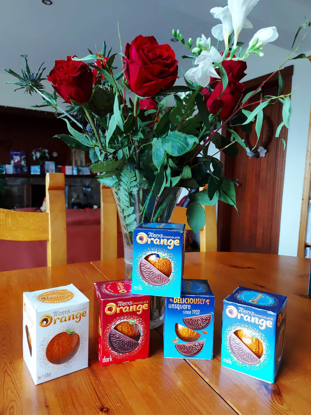 Five Terry's Chocolate Oranges lined up together on a wooden table in front of a vase of Winter flowers. The boxes are different colours to indicate the contents - white, dark and milk chocolate