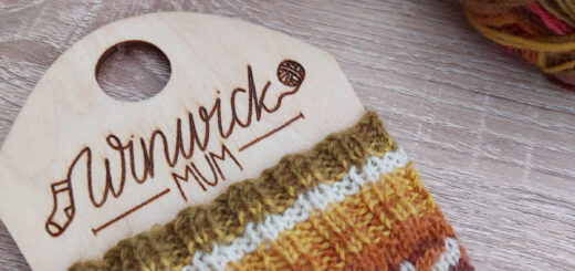 A partly-knitted sock in Autumn shades of brown, green and red with a cream contrast yarn is on a sock blocker with the words Winwick Mum lasered into the wood