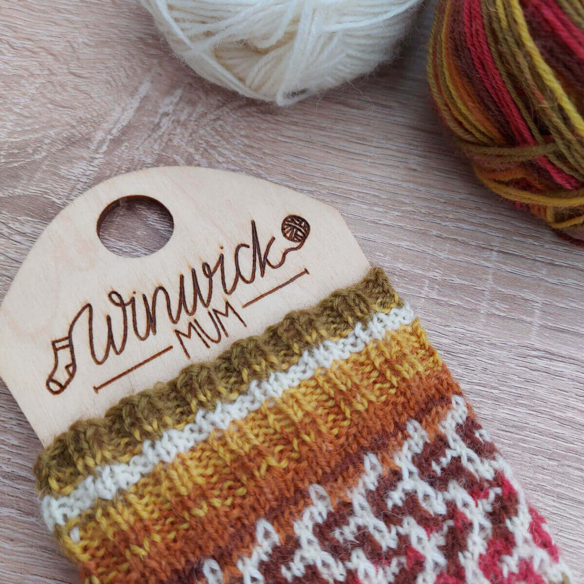 A partly-knitted sock in Autumn shades of brown, green and red with a cream contrast yarn is on a sock blocker with the words Winwick Mum lasered into the wood