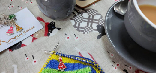 A green, blue and yellow striped sock with a red Christmas tree marker sits on a table next to a grey cup of tea, a grey pottery milk jug, a tea pot and a small card with more stitch markers attached