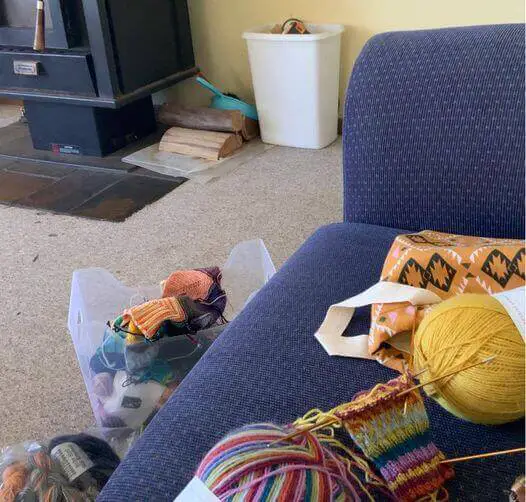 A project bag, balls of yarn and knitting on a blue sofa. Next to the sofa are bags of yarn and in the background, a wood burning stove