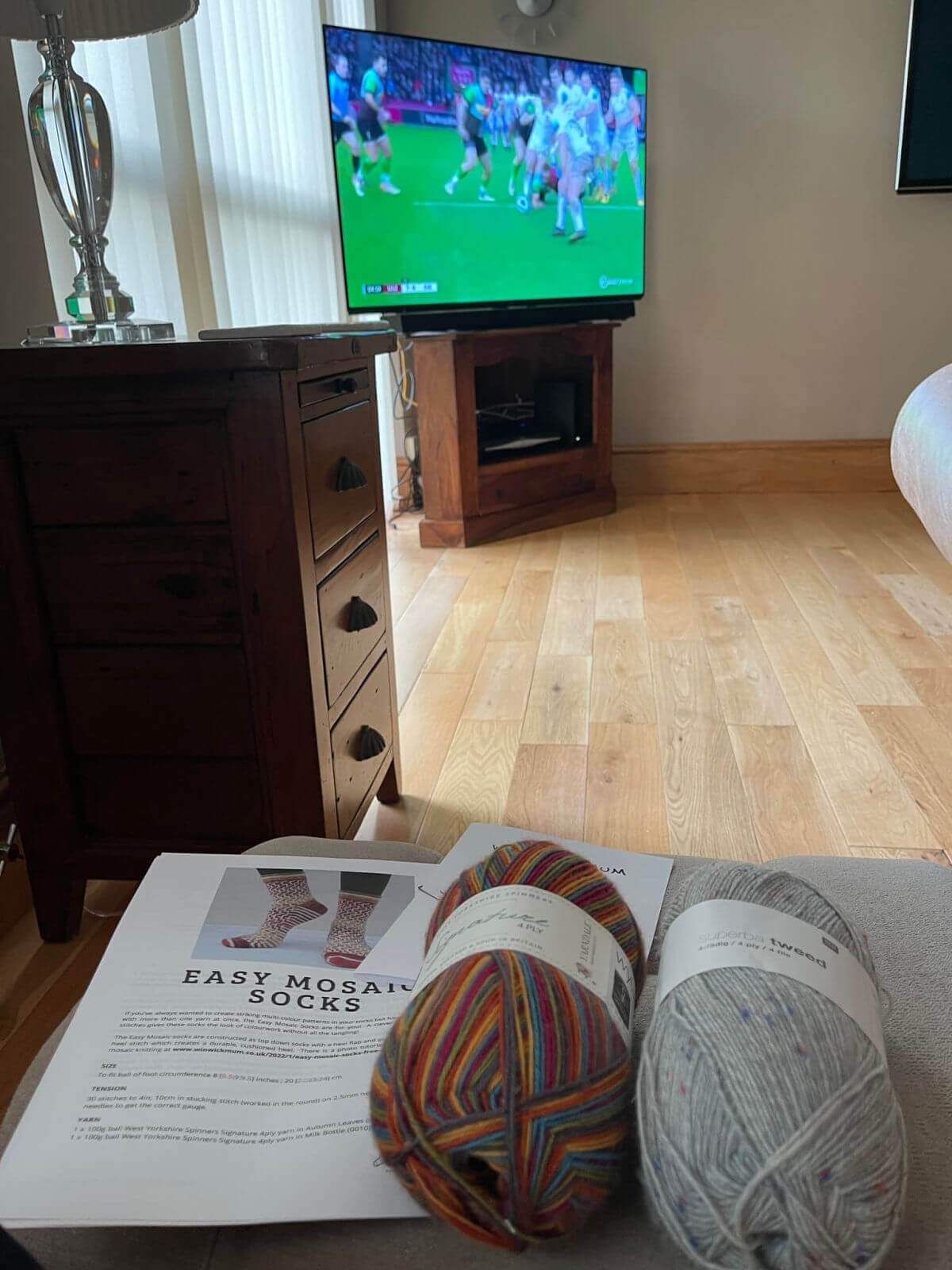 Two balls of yarn are sitting on an Easy Mosaic Socks knitting pattern. In the background is a TV showing a rugby match