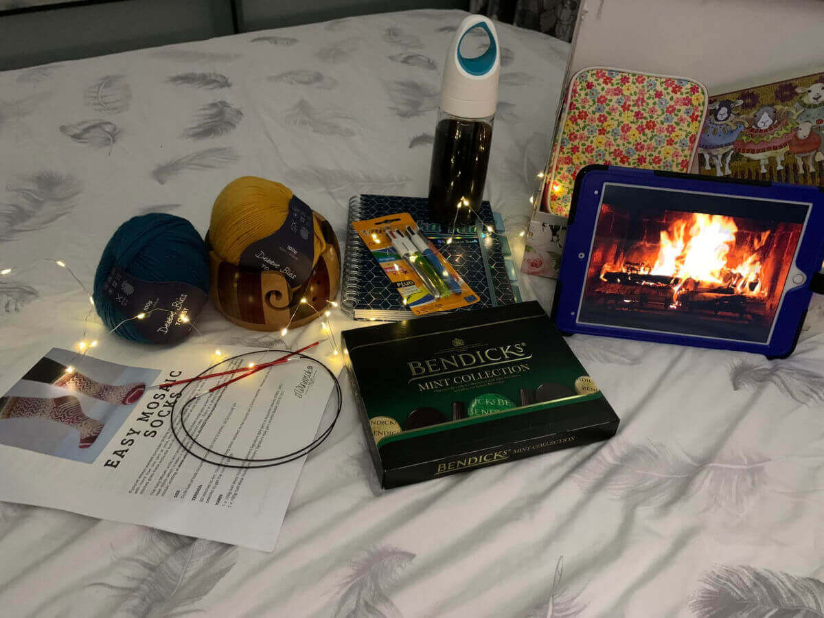 A variety of knitting accessories, yarn, chocolate and an image of a roaring fire on an iPad arranged on a bed on a white duvet cover