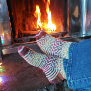 A pair of feet wearing a pair of mosic patterned socks stretched out on a hearth by an open fire. Th