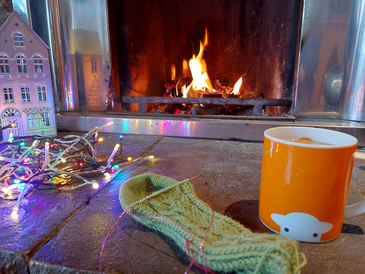 A half-knitted green sock on a stone hearth next to an orange mug of tea. In the background are some coloured fairy lights, a metal house ornament lit up from the inside and a flickering open fire