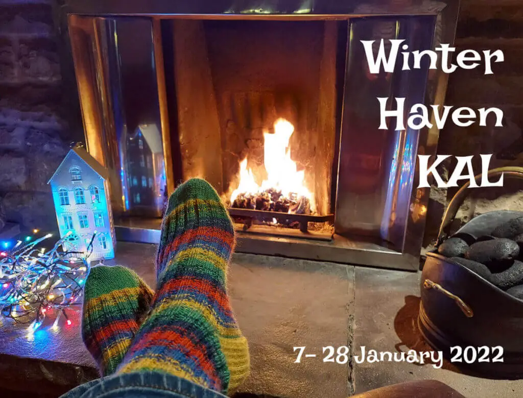 A pair of feet in green, blue, yellow and red striped socks are resting, ankles crossed, in front of a roaring open fire. To the right is a bucket of coal and to the left is an ornamental house decoration, lit up from the inside so that the windows are illuminated, next to a tangle of coloured fairy lights