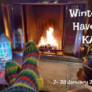 A pair of feet in green, blue, yellow and red striped socks are resting, ankles crossed, in front of a roaring open fire. To the right is a bucket of coal and to the left is an ornamental house decoration, lit up from the inside so that the windows are illuminated, next to a tangle of coloured fairy lights