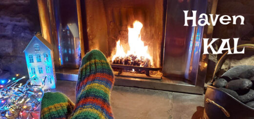 A pair of feet in green, blue, yellow and red striped socks are resting, ankles crossed, in front of a roaring open fire. To the right is a bucket of coal and to the left is an ornamental house decoration, lit up from the inside so that the windows are illuminated, next to a tangle of coloured fairy lights