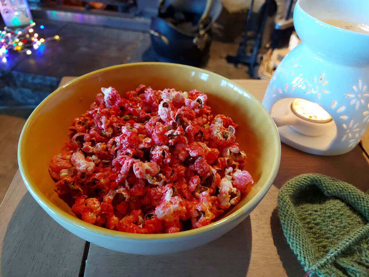 A dish of bright pink popcorn on a coffee table next to a half-knitted green sock, a wax melt burner and in front of an open fire