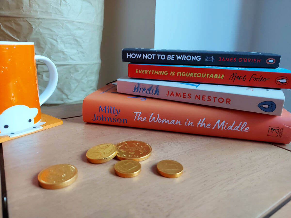 A stack of four books on a light wood coffee table next to an orange mug of tea and five chocolate coins on the table