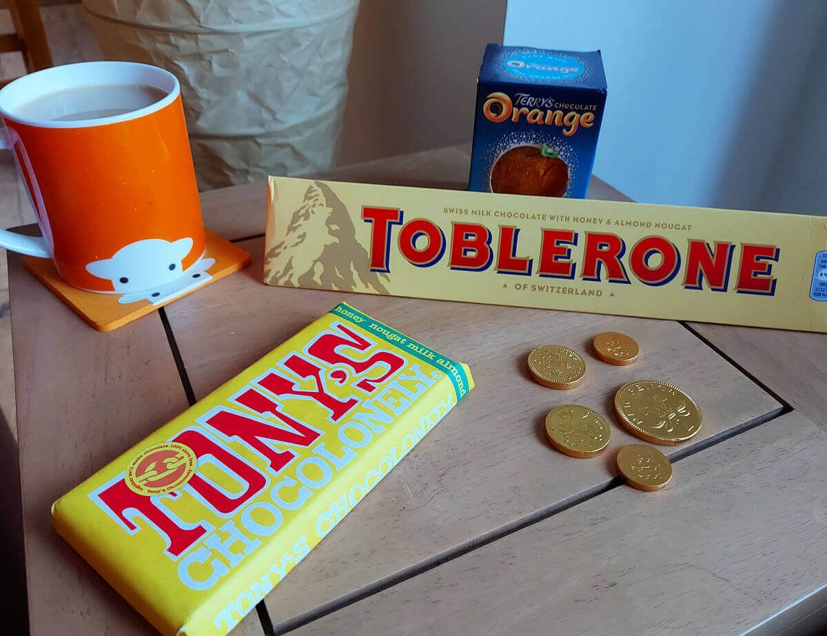 A bar of Toblerone, a chocolate orange, a bar of Tony's Chocolonely chocolate, five chocolate coins and an orange mug of tea on a light wood table.