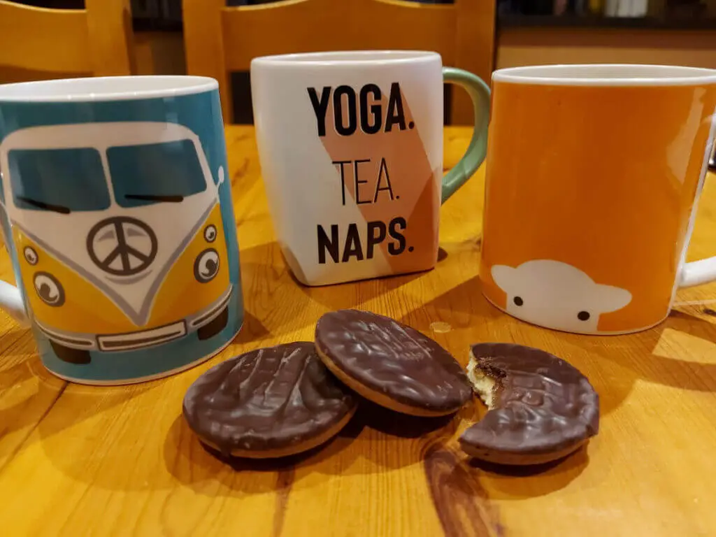 Three mugs on a wooden dining table - left is a turquoise mug with a camper van on it, middle is large white mug with an orange band and the words Yoga, Tea, Naps, and the third is an orange mug with a white sheep's face on it. In front of the mug are three round chocolate biscuits