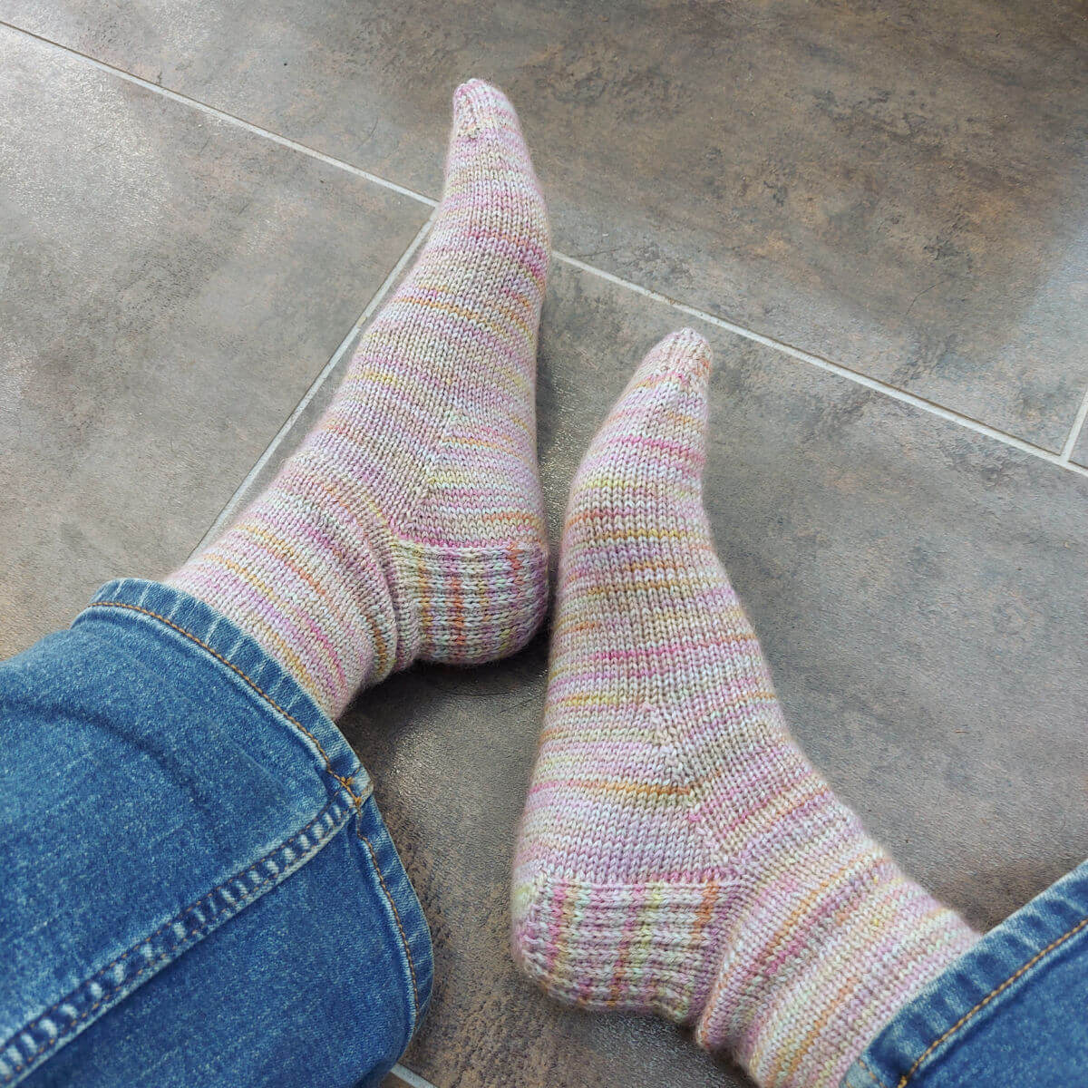 Christine is sitting on a brown and copper-coloured tiled floor showing off a pair of hand knitted socks in pastel shades that give an overall purple tone. She's also wearing her jeans.